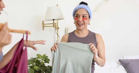Smiling woman comparing wardrobe pieces with friend in bright bedroom casual fashion