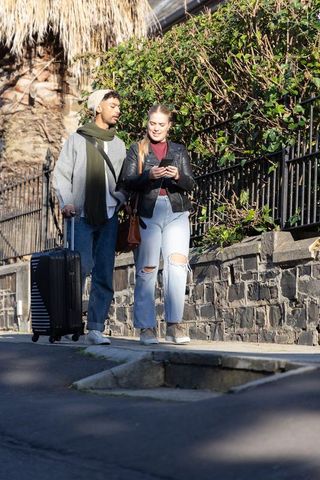 Young couple walking with rolling suitcase while checking smartphone on sunny city sidewalk