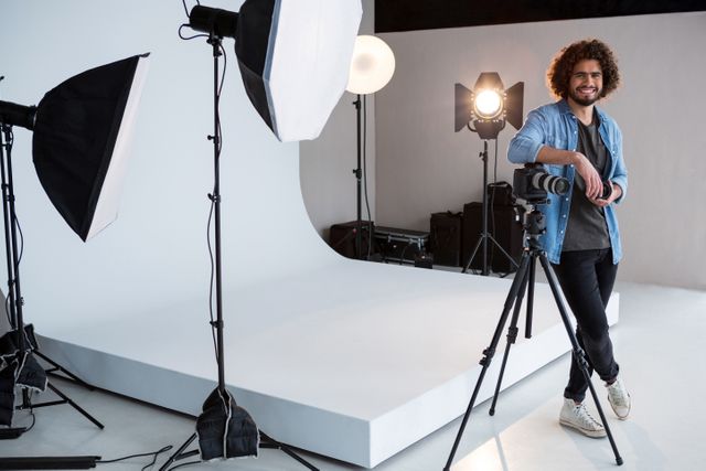 Male photographer standing in a professional studio with camera and lighting equipment. Ideal for use in articles about photography, creative professions, studio setups, and professional workspaces.