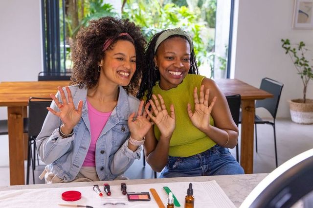 Diverse friends filming joyful at-home makeup tutorial waving at camera with cosmetics