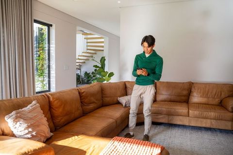 Young asian man standing in modern living room using smartphone near brown leather sofa