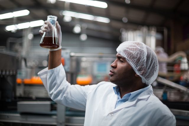 Factory engineer in a white lab coat and hair net is inspecting a liquid sample in a flask at a production plant. This image conveys themes of quality control, professionalism, and industrial processes. It can be used in articles or advertisements related to manufacturing, drinks production, quality assurance, and industrial safety.