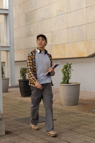 Asian teen male student walking campus plaza holding notebooks wearing beanie and headphones