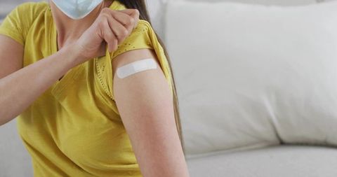 Woman revealing vaccination bandage on upper arm wearing mask in yellow shirt at home