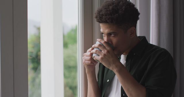 Thoughtful biracial man looking through the window and drinking coffee. quality time, relaxing at home.