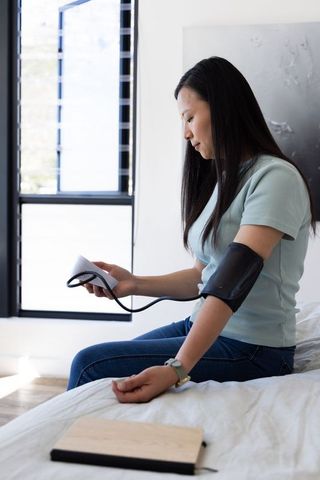 Asian woman measuring blood pressure at home bedroom using digital arm cuff and monitor