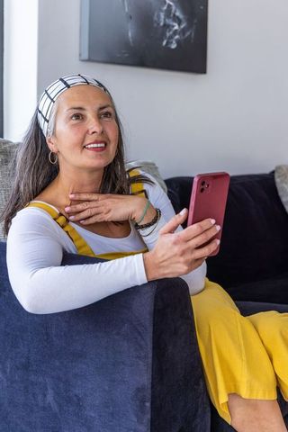 Middle-aged woman smiling on navy sofa holding smartphone wearing yellow dress and headband
