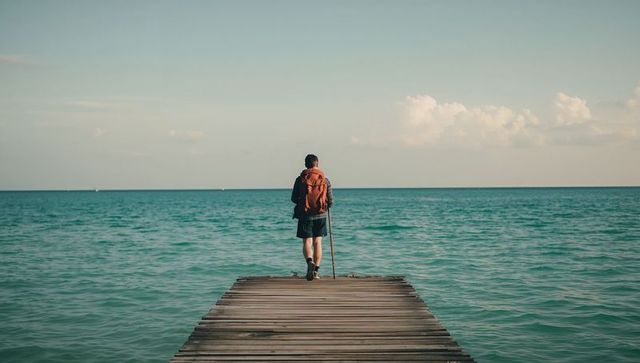 Solo hiker standing on wooden pier facing teal sea with rust backpack and walking pole