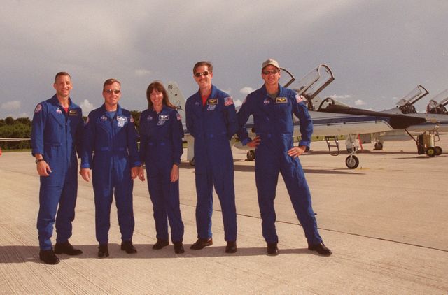 Five astronauts wearing blue flight suits standing on tarmac beside spacecraft at Kennedy Space Center. Ideal for use in articles about space exploration, astronaut training, NASA missions, or team collaborations. This image evokes themes of teamwork, human spaceflight, and technological achievement with connotations of NASA and space exploration programs.