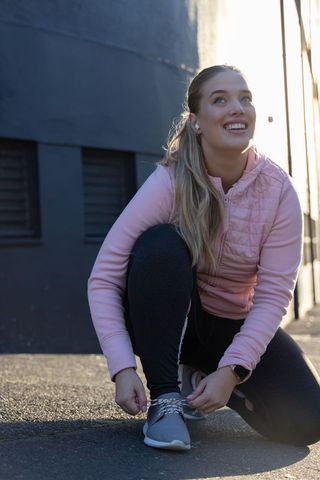 Smiling woman tying running shoe in sunlit urban alley wearing pink jacket and smartwatch