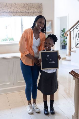 African american mother supporting daughter holding 1st day of school letterboard in home entry