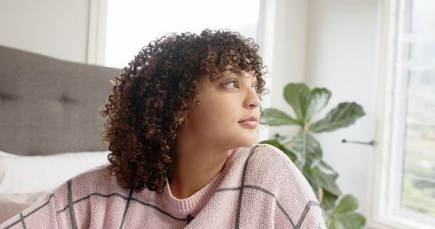 Young woman wearing pink sweater gazing out window in serene bedroom with fiddle leaf fig