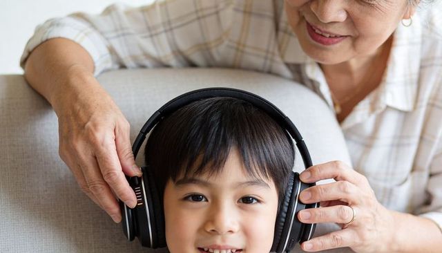 Grandmother adjusting headphones on smiling child on sofa, intergenerational family bonding