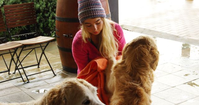 A young woman with blonde hair wearing a beanie and casual clothes dries her two golden retrievers with an orange towel under a shelter during a rainy day. A barrel and a wooden bench are visible in the background. This image is suitable for content related to pet care, animal love, bonding moments with pets, dog breeding, dog lover blogs, and advertisements for pet grooming products.