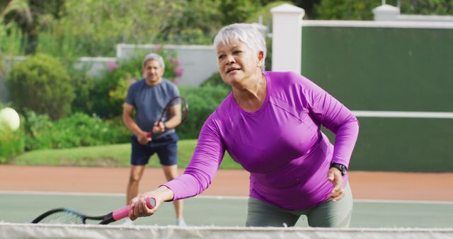 Image of happy biracial senior woman practicing tennis on tennis court. active retirement lifestyle, senior relationship and tennis training concept.
