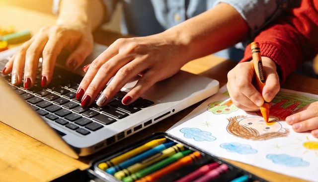 Parent multitasking typing on laptop while child coloring with crayons family creativity