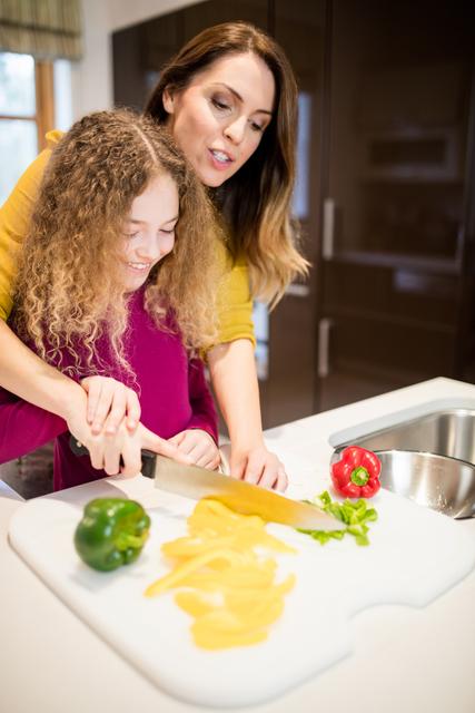 Image depicts mother assisting her daughter with cutting vegetables on a white cutting board in a modern kitchen setting. Suitable for use in themes of family bonding, cooking tutorials, healthy eating, home activities, and parenting articles.