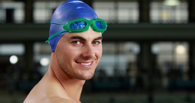Male swimmer wearing blue swim cap and green goggles, smiling confidently. Ideal for use in content related to swimming, professional athletes, competitive sports, aquatic activities, health and fitness.