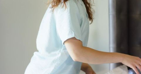 Leaning woman adjusting white bed linens in bright bedroom, closeup torso and arm