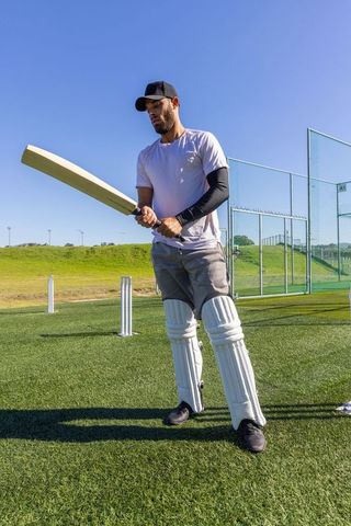 Cricketer practicing batting on synthetic pitch wearing pads and cap in sunny outdoor nets