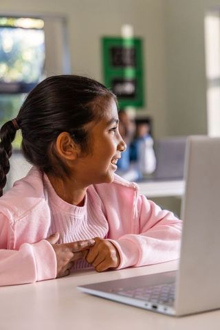 Elementary girl smiling while learning on laptop in bright classroom, focused engagement