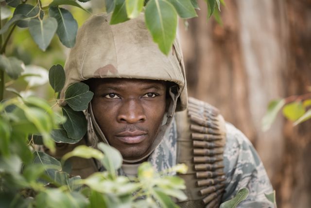 Military soldier in camouflage uniform and helmet hiding behind trees during boot camp training. Ideal for use in articles about military training, combat readiness, tactical exercises, and survival skills. Can also be used in promotional materials for military recruitment and defense strategies.