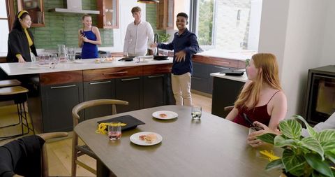 Diverse young adults enjoying casual kitchen gathering with smartphones, pastries and drinks