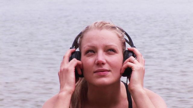 A young woman is relaxing by the beach while listening to music on her headphones, embodying a sense of tranquility and leisure. This visual is ideal for themes related to self-care, music enthusiasts, travel, relaxation, and lifestyle marketing materials. It captures a perfect moment of escape and personal enjoyment.