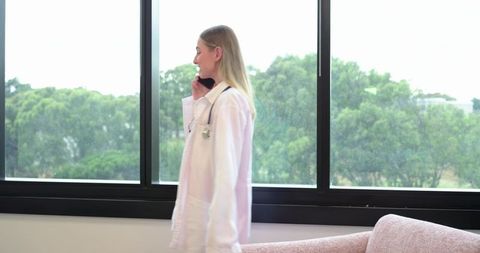 Female doctor walking past window talking on phone wearing lab coat and stethoscope