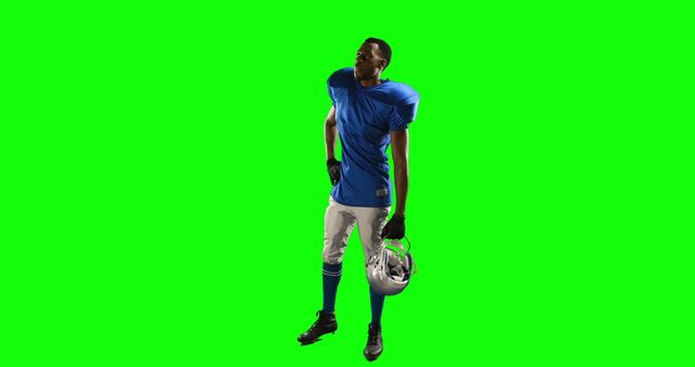 American football player standing wearing blue jersey, holding helmet, posing against green screen background. Great for use in sports advertisements, promotional materials for football events, and creating composite images for media projects.