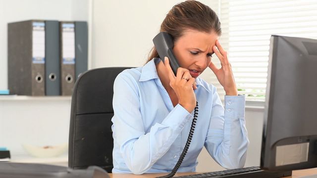 The image shows a woman experiencing stress at her workplace while ending a phone call, with documents and a computer present. Useful for illustrating themes related to workplace stress, business communication challenges, and stress management techniques.