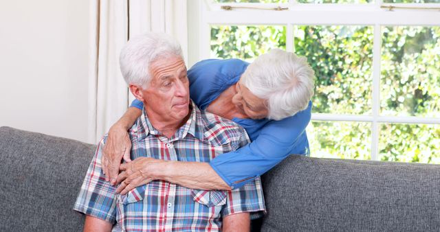 Elderly couple sharing a loving embrace on a sofa at home, exuding warmth and affection. This image is perfect for content related to senior living, family bonds, retirement, and emotional well-being in later stages of life. It can be used on websites, blogs, advertisements, and brochures focusing on elderly care, family relationships, and home living environments.