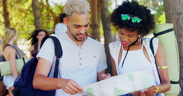 Young couple with backpacks is standing outdoors in a forest, studying a map intently. They are dressed casually in summer clothing, with bright daylight filtering through the trees. This can be used for topics such as travel, adventure, hiking, exploring nature, or promoting eco-tourism.