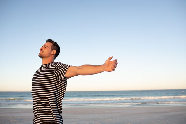 Man standing on beach with arms outstretched, enjoying the sunset. Ideal for themes of relaxation, freedom, mindfulness, and nature. Perfect for travel blogs, wellness articles, and advertisements promoting vacations or mental well-being.