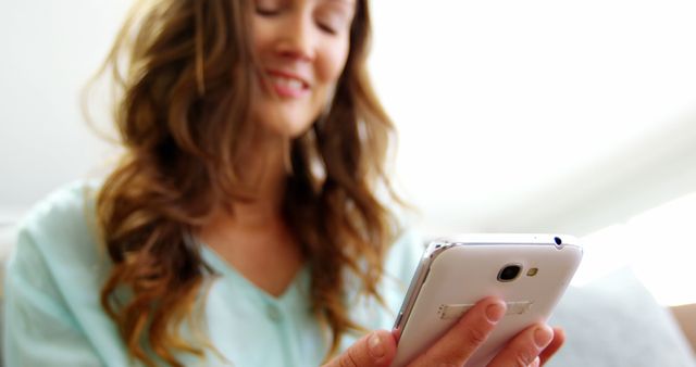 Smiling woman comfortably using smartphone indoor provides a relatable scene for promoting mobile apps, social media engagement, communication services, or lifestyle content. Useful for marketing campaigns focused on tech-savvy lifestyles or for editorial content discussing the integration of technology in daily lives.