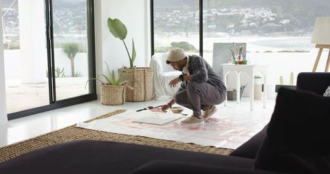 Indian artist painting on floor in bright coastal studio wearing bucket hat