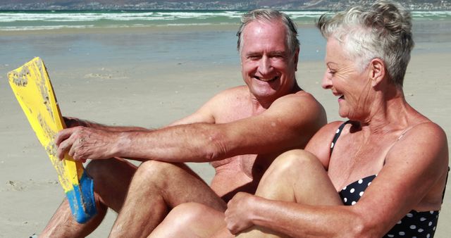 A senior Caucasian couple enjoys a playful moment on the beach, with the man holding a yellow paddleboard and both smiling broadly. Their joyous interaction captures the essence of active and happy retirement years.