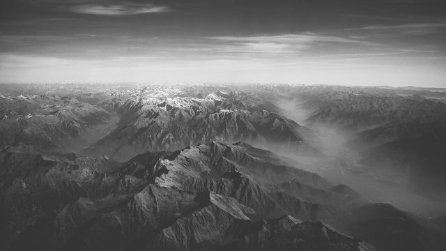 Spectacular bird's-eye view of an expansive mountain range with dramatic lighting casting shadows over the rugged terrain. The monochromatic black and white filter emphasizes the contrast and texture of the mountains, adding a timeless quality. Ideal for use in travel brochures, nature photography collections, inspirational posters, and environmental campaigns.