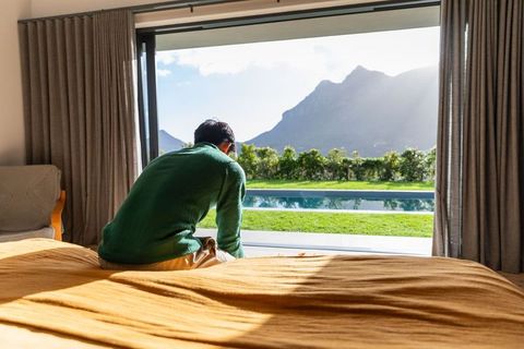 Man sitting on bed with mustard duvet, sliding doors opening to pool and mountain view