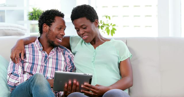Two African American adults are sitting closely on a couch, sharing a moment while looking at a tablet, with copy space. Their relaxed posture and smiles suggest a comfortable and happy interaction, enjoying leisure time together.