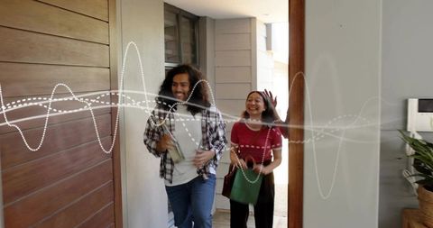 Young couple entering home carrying groceries and green tote smiling with digital waveform overlay