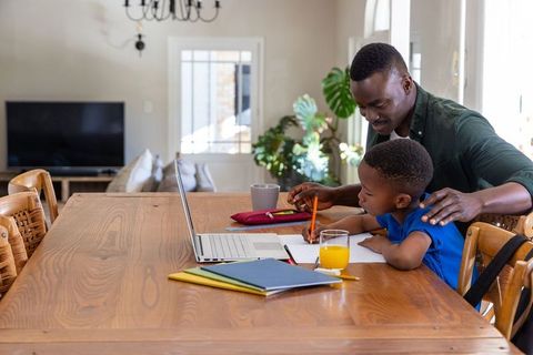 African american father guiding son through homework at wooden dining table with laptop