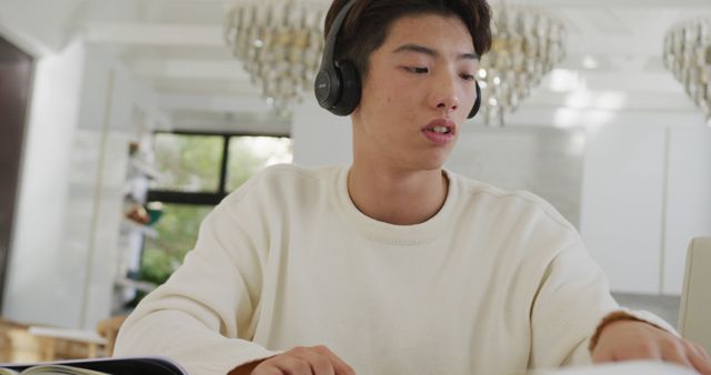 Asian male teenager with headphones learning and using laptop in living room. spending time alone at home.