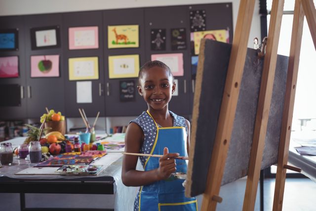 Young girl is painting on a canvas in an art classroom, smiling at the camera. The classroom is filled with various art supplies and colorful artworks displayed on the walls. Ideal for use in educational materials, art class promotions, children's creativity and learning resources, and school brochures.