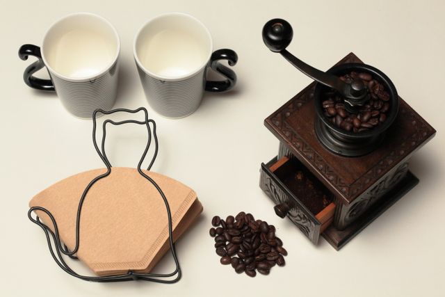 Old-fashioned coffee grinder filled with coffee beans, two coffee cups, and brown coffee filter on clean, neutral background. Ideal for illustrating morning coffee rituals, coffee lover themes, retro kitchen decor, and barista tools. Perfect for lifestyle blogs, coffee shop promotions, and vintage kitchen design inspiration.