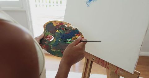 African american woman painting on canvas at easel with colorful palette and brush