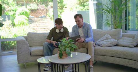 Father and teen son sharing tablet while gaming on sofa in sunlit modern living room