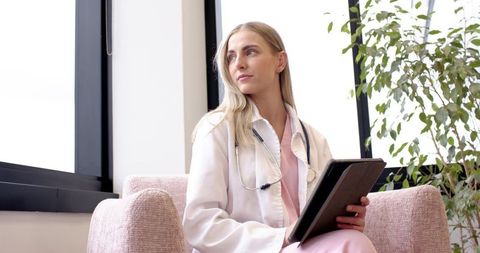Female doctor holding tablet gazing out window in sunlit clinic waiting area