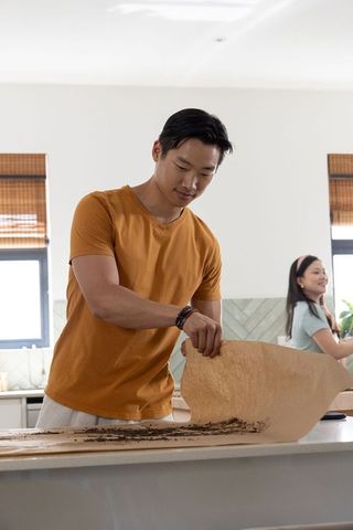 Asian couple cooking together in sunlit modern kitchen man lifting parchment with crumbs