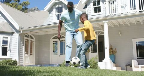 African american father playing soccer with son on front lawn of white suburban house